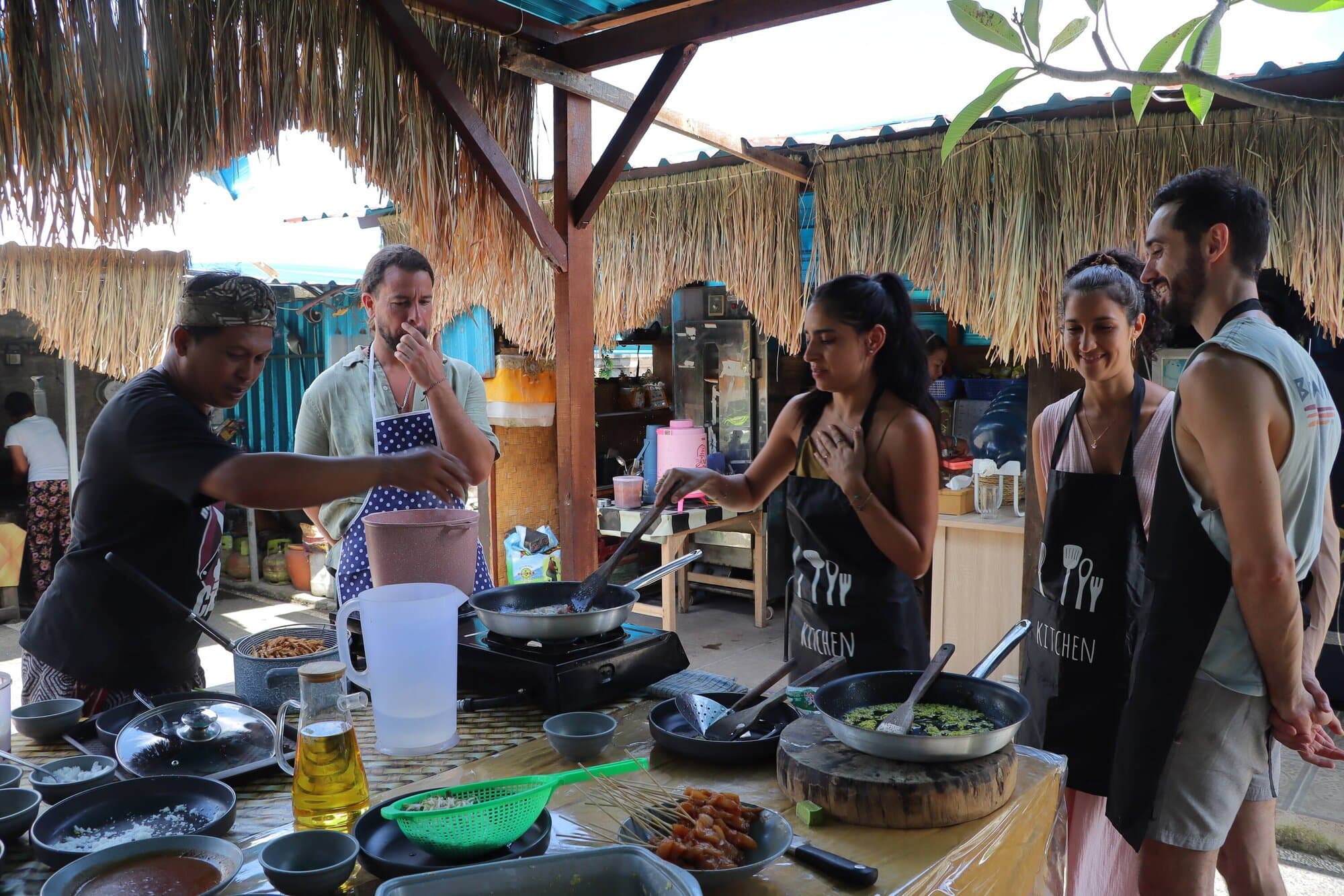 Tribe members in aprons cooking together with Dewa under a thatched warung roof.